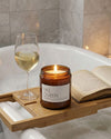 Candle labeled 'YAS QUEEN' on a bathtub tray with a glass of wine and an open book.