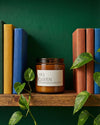 Candle labeled 'YAS QUEEN' on a wooden shelf with books and greenery against a green wall.